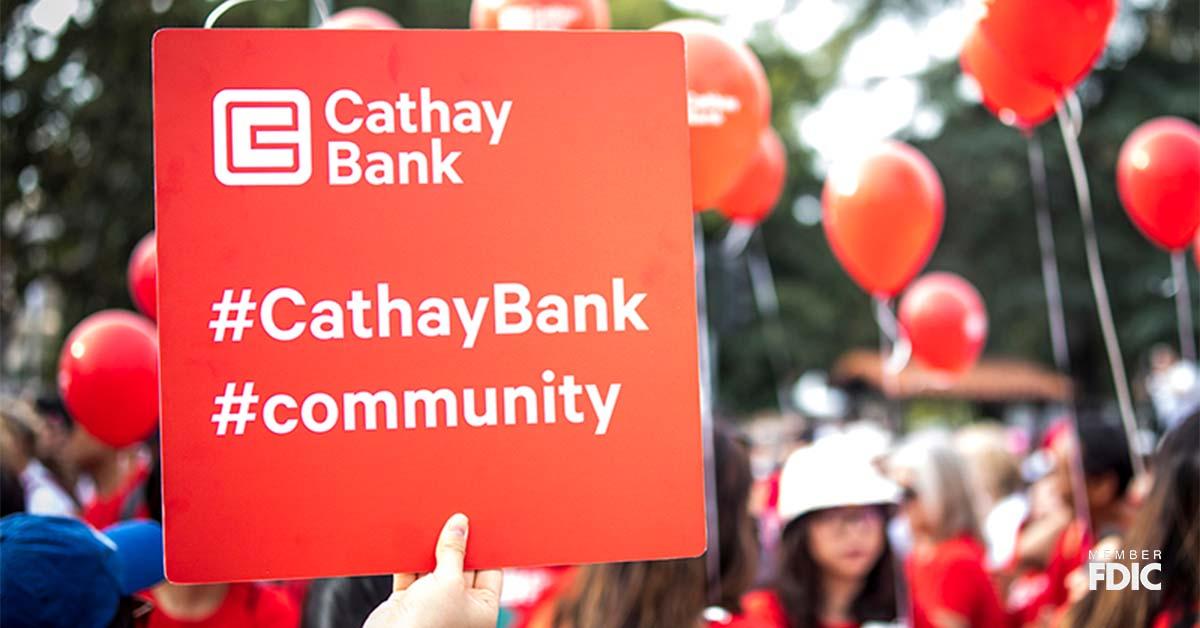 A red sign is being held by a person in a crowd which shows the Cathay Bank logo as well as hashtags for community and Cathay Bank