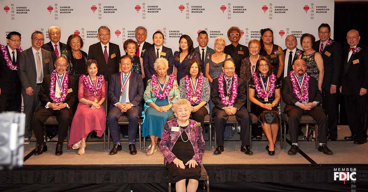 Un grupo de personas se sientan en la primera fila y se paran en la última fila en un escenario como invitados especiales y homenajeados para la Gala de Historiadores del Museo Chino Estadounidense en Los Ángeles.