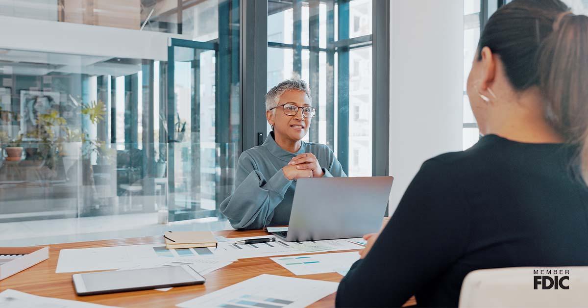 A business client sits while facing an auditing advisor inside a bright office while they review paperwork over a wooden desk.