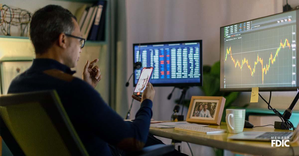 A businessman holds his cellphone to video chat a client overseas while sitting in front of his desk, viewing the foreign exchange stats on the computer screen.