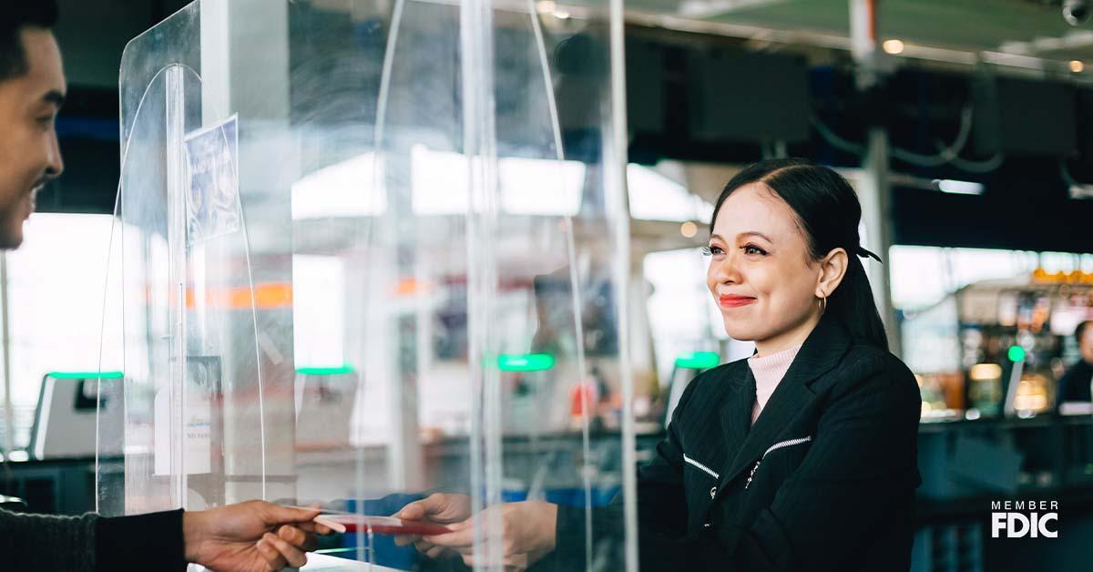 A bank teller stands behind a protective glass shield while handing a checkbook back to a client.