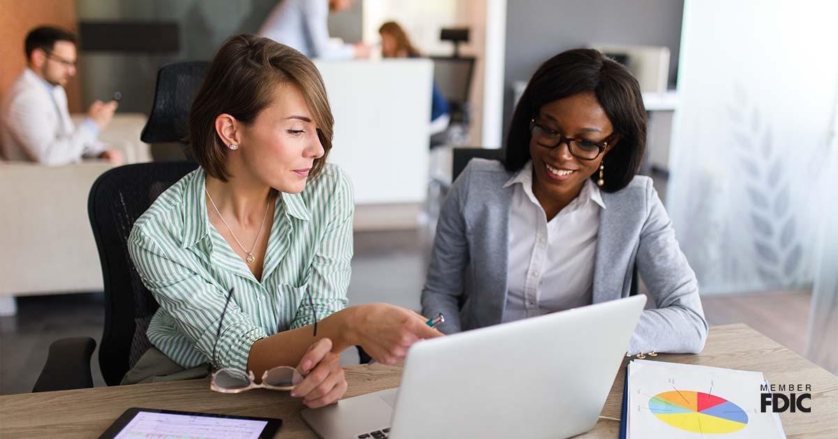 Two business women chat over international trade finance while looking at a laptop in an office.