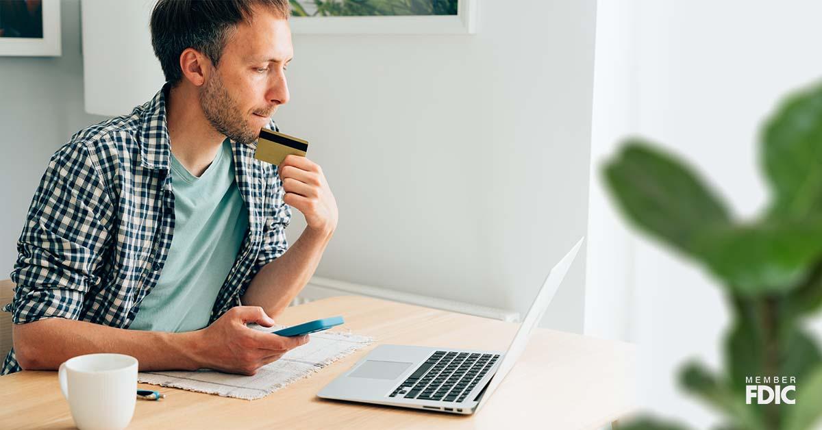 A man sits at his wooden desk in a brightly lit office while staring at his laptop and holding his credit card with a worried look about fraud alerts