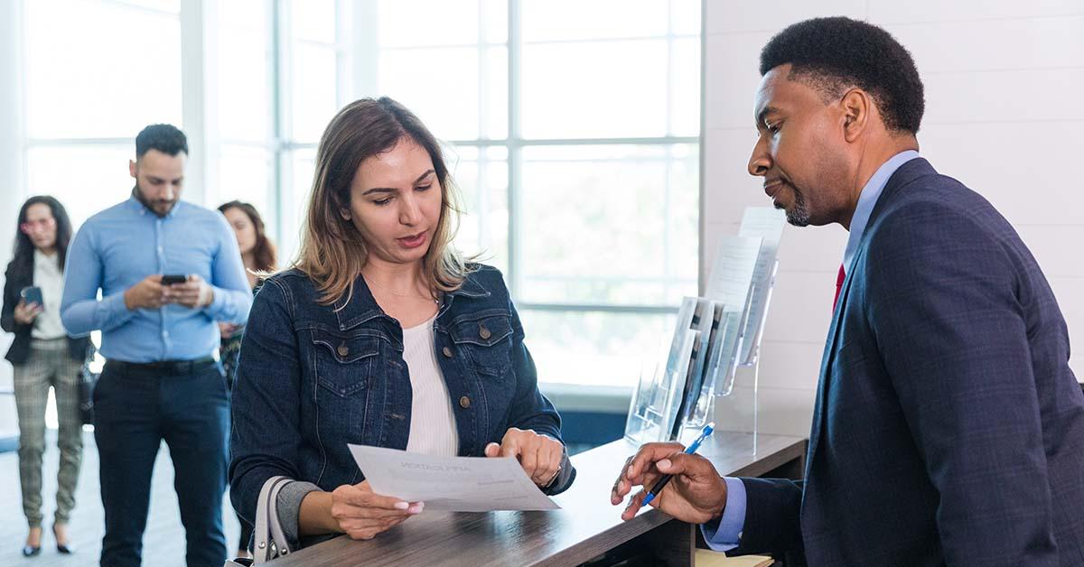 An adult male bank manager stands at the counter to answer the adult woman's questions about her monthly statements and recent check cashing.