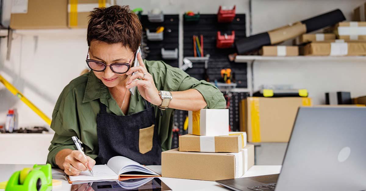 An employee at a small business wearing a green blouse with black overalls smiles as she speaks with a customer on the phone over her office desk.