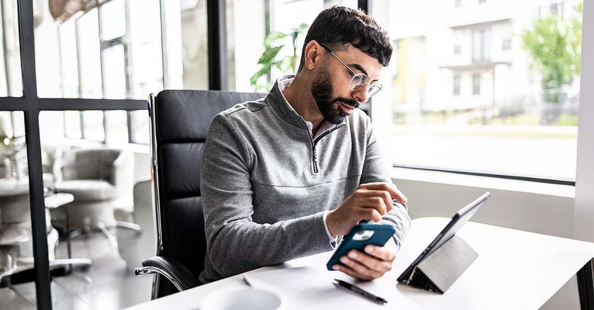 Un hombre con un suéter gris y gafas está sentado en su escritorio. En la mano izquierda sostiene un teléfono, mientras que con la derecha apunta a un iPad que está sobre el escritorio frente a él.