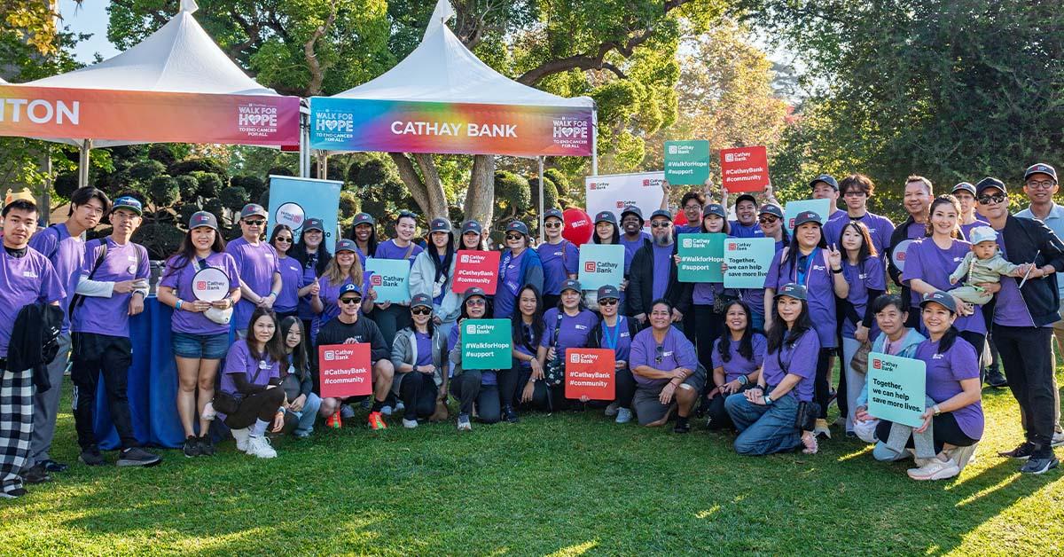Cathay Bank team members in purple t-shirts stand together in an outdoor grass area with a Cathay Bank tent behind them, holding Cathay Bank signs for a group photo.