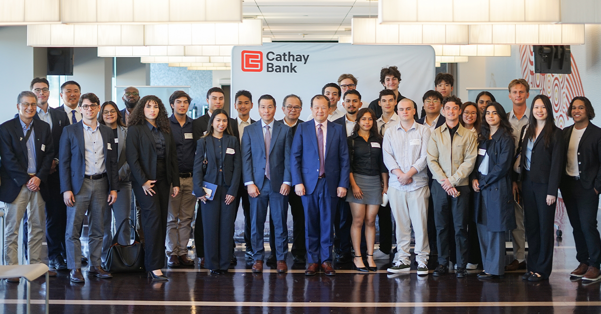 Un numeroso grupo de estudiantes y representantes de Cathay Bank posan juntos frente a un fondo con el logotipo de Cathay Bank. El grupo viste ropa formal o semiformal de negocios y posa para una foto grupal formal dentro de las oficinas corporativas, con iluminación y decoración interior modernas de fondo.