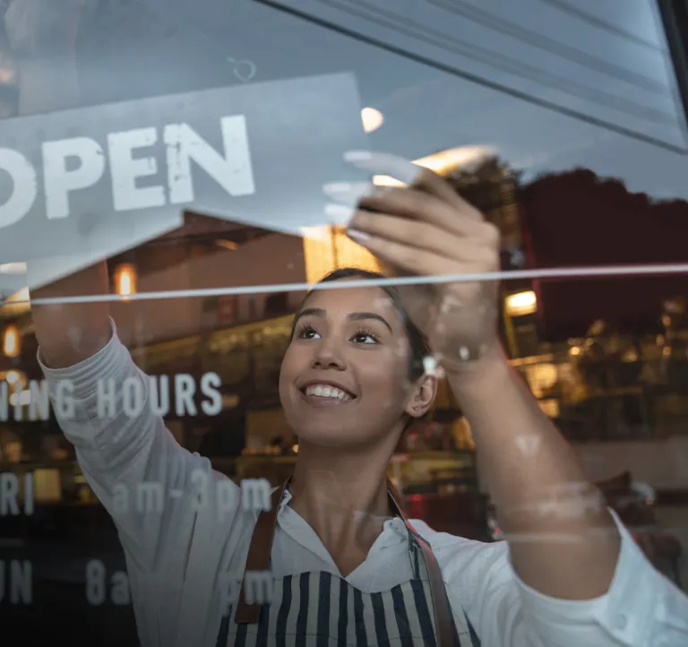 A small business owner adjusts an “open” sign on the front window of a shop. Reflections of the street, cars, and warm interior lights show a lively neighborhood environment.