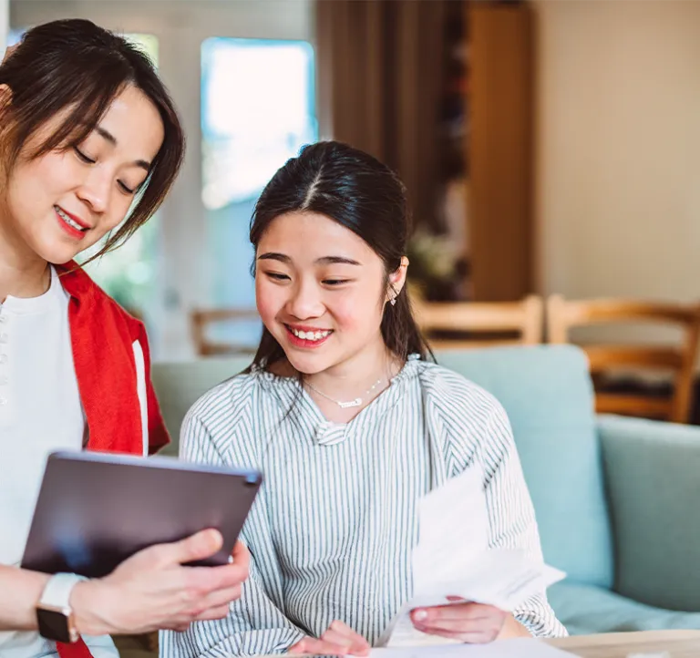 Two women sit on a couch, smiling as they review information on a tablet and a printed document in a bright living room.