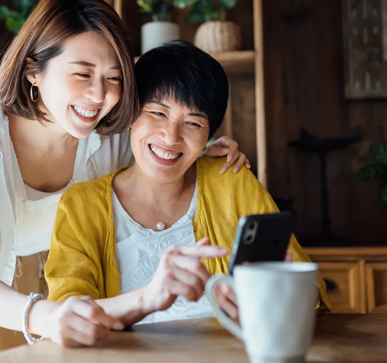 Two people indoors, standing close together and looking at a smartphone. One person is seated holding the phone while the other stands slightly behind with an arm resting on the seated person’s shoulder. Wooden shelving and potted plants are in the background