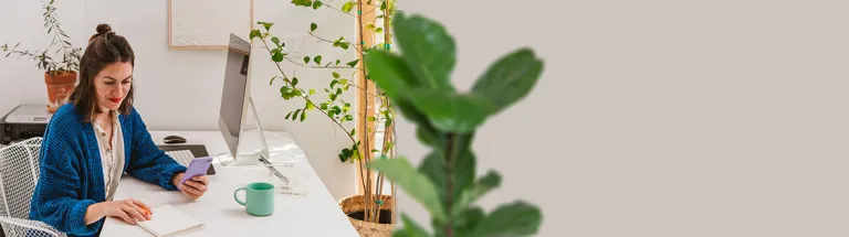 A woman sits at her desk while planning for finances with Zelle for Business and holding her mobile phone in a bright lit office with various green plants.