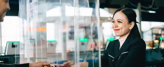 A bank teller stands behind a protective glass shield while handing a checkbook back to a client.