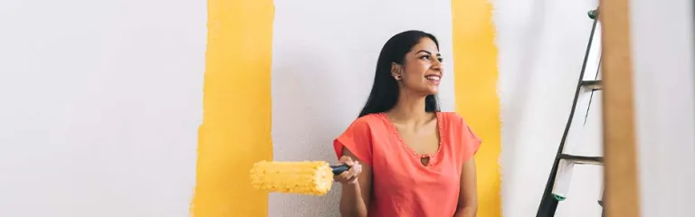 A smiling young woman with paint roller sitting by ladder at home