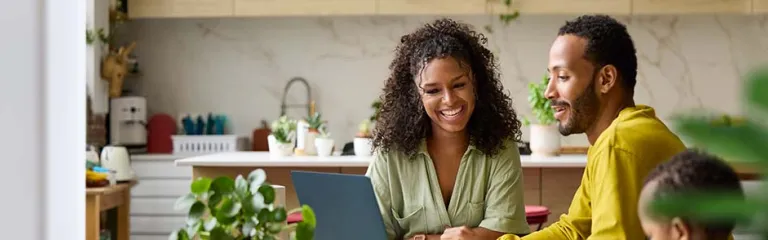 Freelancer discussing with smiling woman over laptop. Parents are sitting by toddler boy drawing at table. They are at home.