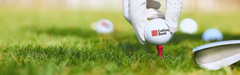 A golf ball with Cathay Bank logo sits outdoors on grass with a golf club and hand next to it for the annual tournament in California. 