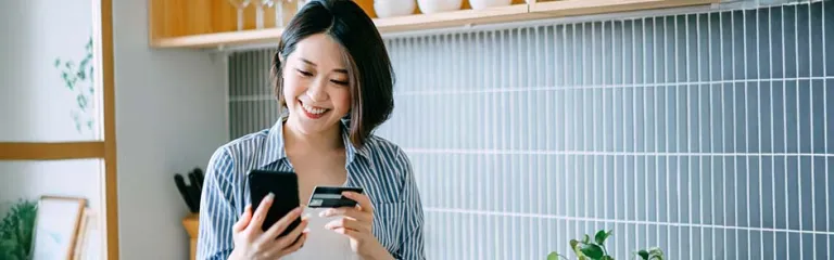 A woman is standing in a modern kitchen holding a smartphone in one hand and a credit card in the other, suggesting an online payment or mobile banking activity. 