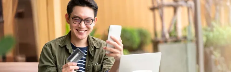 A man is sitting at a table outdoors with a laptop, holding a smartphone in one hand and a credit card in the other, suggesting an online payment or mobile banking activity. 