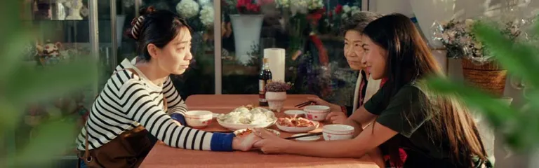 Family sharing a meal at a table with traditional dishes, holding hands in celebration of Lunar New Year, surrounded by flowers and warm décor.
